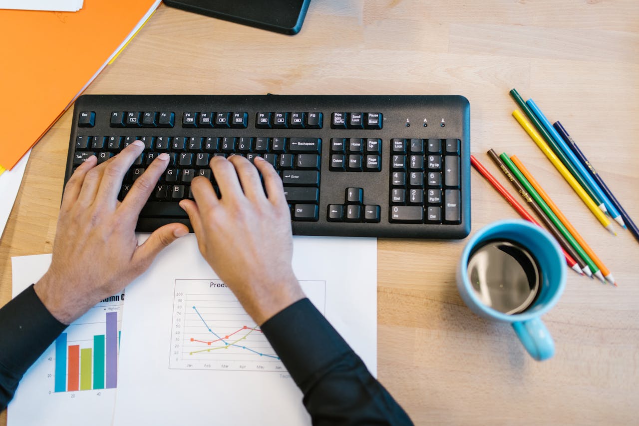 Workspace featuring hands typing on a keyboard, a coffee mug, colored pencils, and graphs on paper.